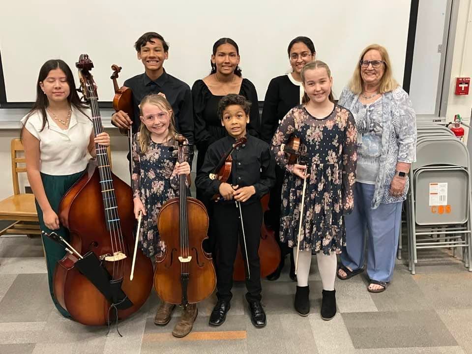 Six students of varying ages holding a double bass, cello and violins, standing with their teacher.