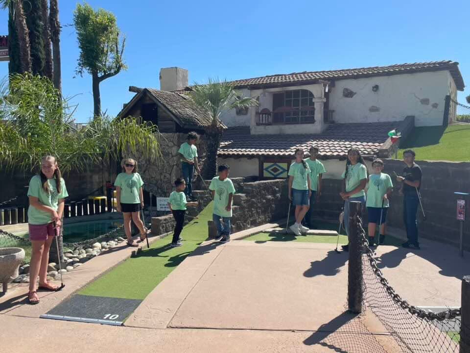 Students in matching studio t-shirts playing a round of miniature golf together on a sunny day.